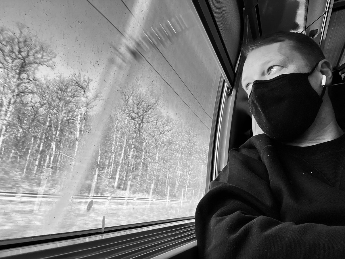 A black and white photography of a man wearing headphones and a face-mask looks out of the window of a moving train. There are trees outside the window.