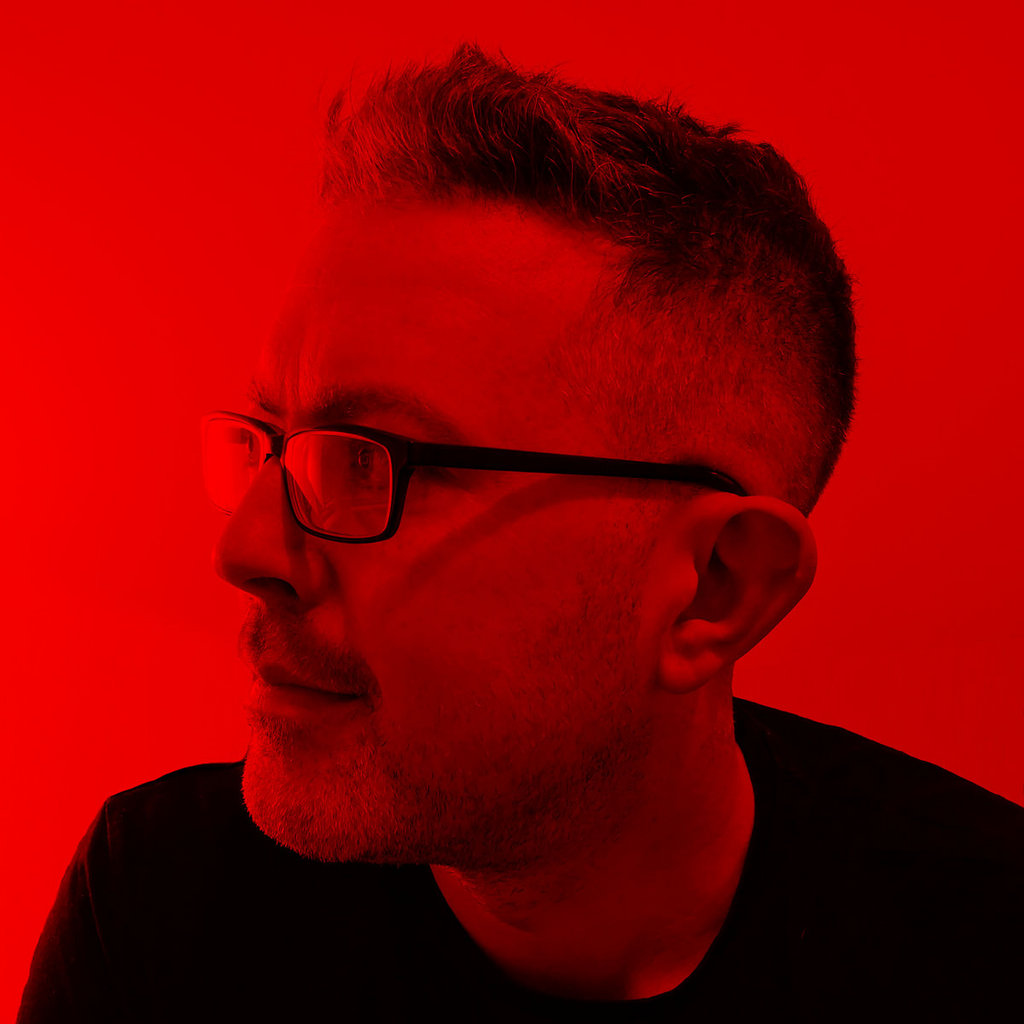 A red-tinted close up photo of a man with short hair and glasses, looking off camera